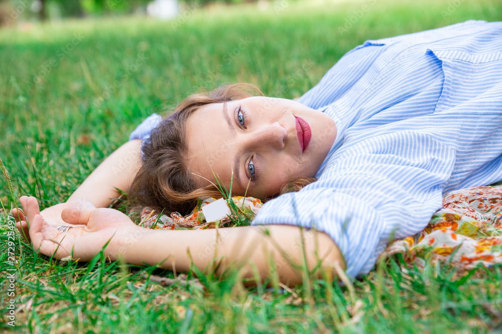 Calm young woman lying on grass and looking at camera. Content girl in shirt relaxing in city park. Laziness concept Calm young woman lying on grass and looking at camera. Content girl in shirt relaxing in city park. Laziness concept
