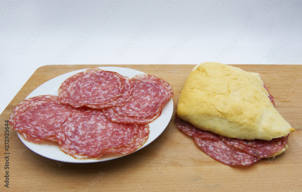 Fresh made salami sandwich, with slices of salami in a ceramic white dish, on wooden background