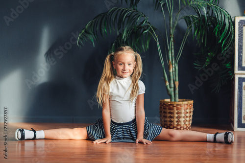 Beautiful little blonde girl with hair gathered in tails, white t-shirt, white socks and gray skirt sitting on twine at home looking at camera and smiles.