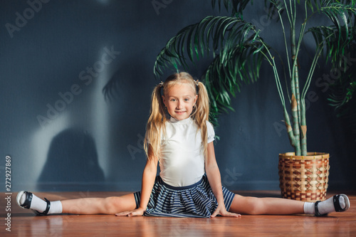 Beautiful little blonde girl with hair gathered in tails, white t-shirt, white socks and gray skirt sitting on twine at home looking at camera and smiles.