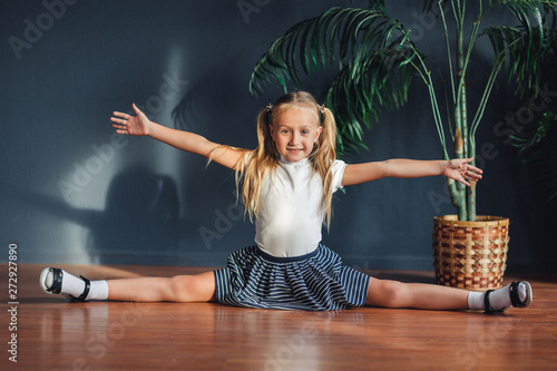 A little girl with hair gathered in tails, white t-shirt, white socks and gray skirt, adorable young talented dancer does ballet poses and stretching exercises on the floor at home