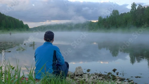 man sits on ground and looks at calm river flow with fog