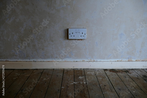 A UK Double Plug Wall Socket, Showing the Power Supply on an Unfinished Interior Wall, with Pine Floor Boards and Staining Remnants.