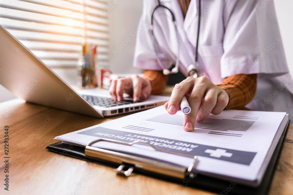 Fototapeta premium Young female in uniform of doctor using digital technology laptop for Output Device and writing a patient report on the office desk in afternoon.