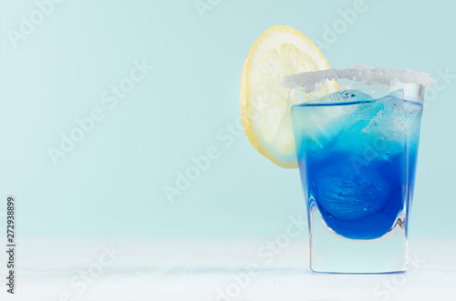 Blue curacao cocktail with lemon slice, ice cubes, sugar rim in shot glass on elegant white wood board and pastel mint color wall.