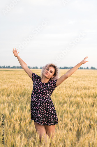 young tanned curly haired woman in dress is happy to be on the field of ripened wheat, sunset time
