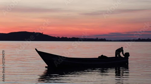 Two young fishermen stand up in a small wooden boat on Lake Victoria under orange evening sky. Silhouettes show how one of them tries to get the engine started. photo