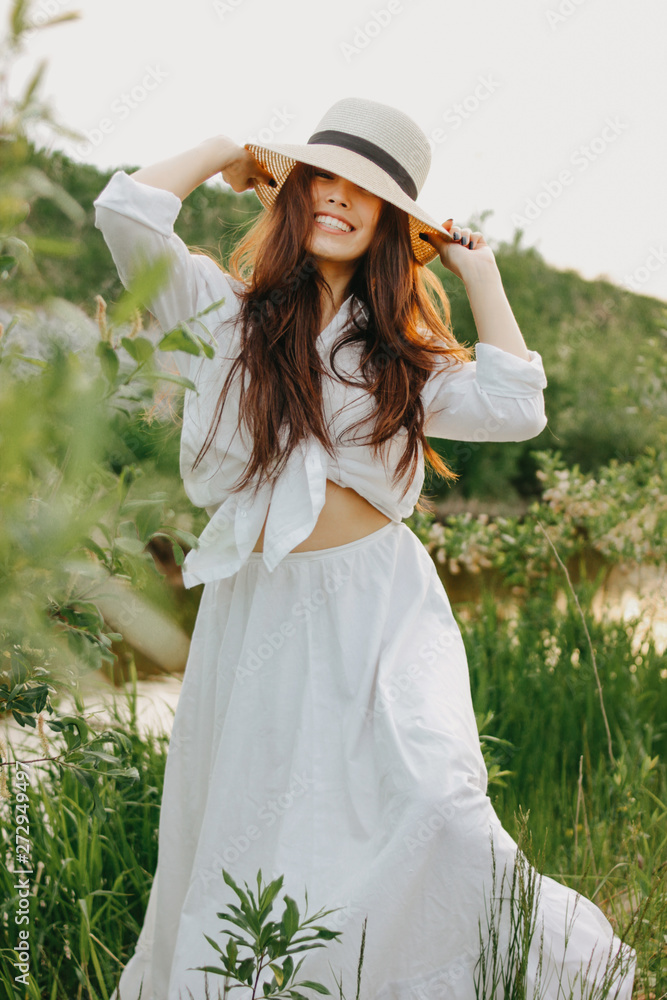 Φωτογραφία Beautiful carefree long hair asian girl in white clothesand straw hat  on river side in the grass at sunset