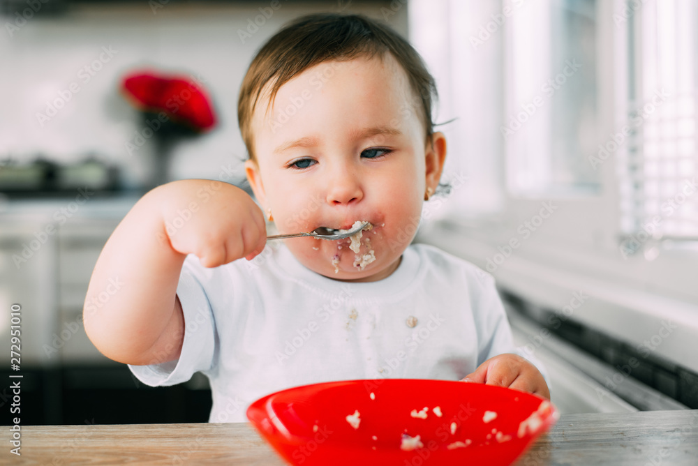 Funny little girl in the kitchen during the day eating oatmeal porridge from a red plate all dirty Funny little girl in the kitchen during the day eating oatmeal porridge from a red plate all dirty