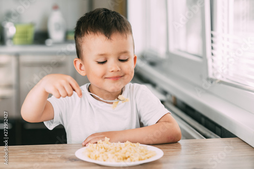 a child in the kitchen during the day eating pasta in a spiral in a white t-shirt