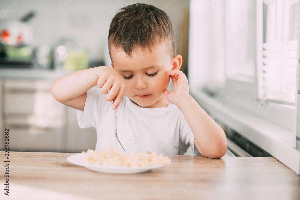 Fototapeta premium a child in the kitchen during the day eating pasta in a spiral in a white t-shirt