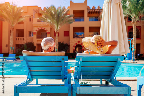 Senior couple relaxing by swimming pool lying on chaise-longues. People enjoying summer vacation.