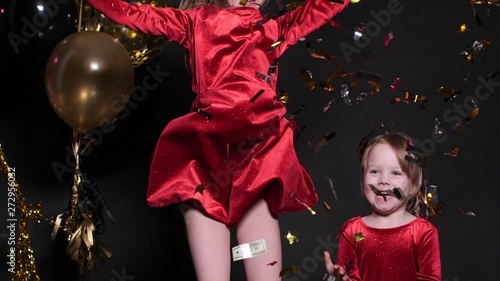 Studio portrait of adorable fair-haired young girl in blue jersey and skirt jumping and having fun with golden sparkling confetti and air balloons on black background. She is happy and cheerful