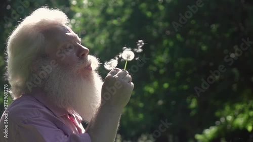 Close up portrait of handsome bearded senior man blowing on dandelions. Fluffy white seed flying away. Beautiful slow motion shot at nature against dark green background.