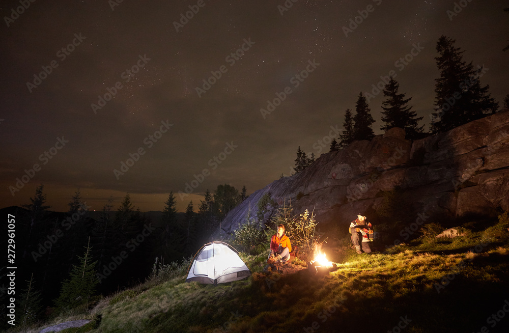 Night camping in the mountains. Young couple hikers having a rest together, sitting beside campfire and illuminated tourist tent. On background big boulder, forest and beautiful night starry sky. - obrazy, fototapety, plakaty 