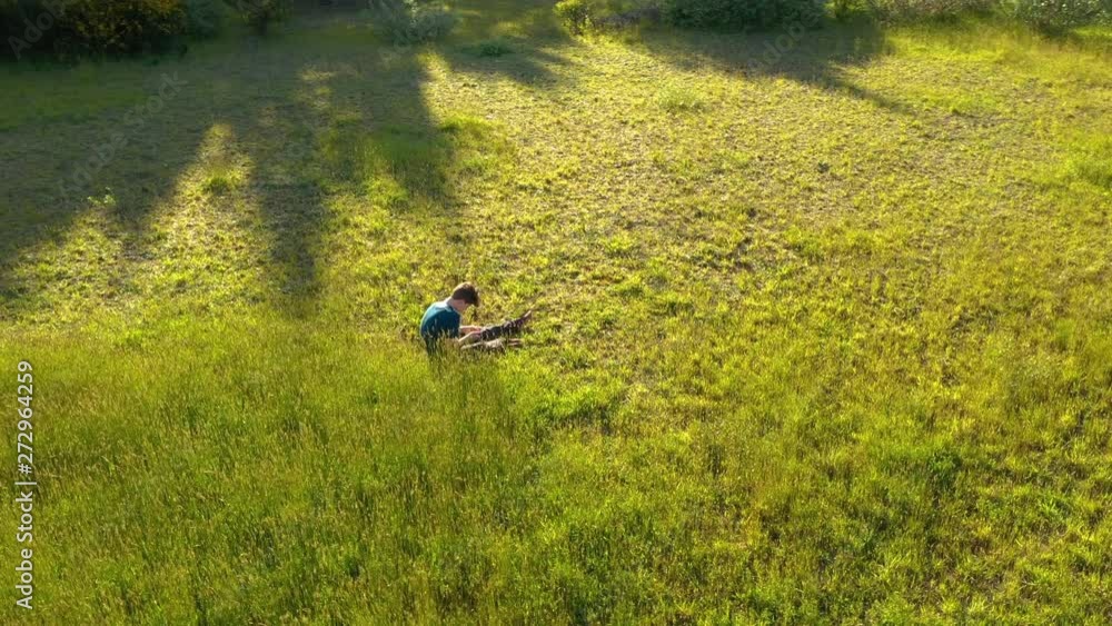 Teenage boy sitting in grass and looking at his smart phone. Drone slowly circling him. Teenage boy sitting in grass and looking at his smart phone. Drone slowly circling him.
