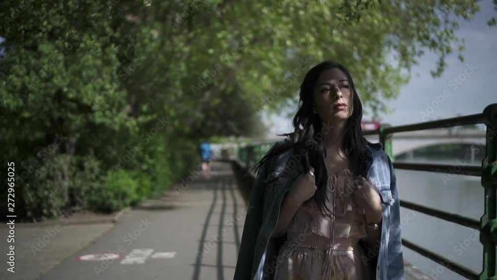 Slow Motion of an attractive latina tourist with black wavy hair and a jean jacket walking in a park in London with a view of Putney bridge behind her Slow Motion of an attractive latina tourist with black wavy hair and a jean jacket walking in a park in London with a view of Putney bridge behind her