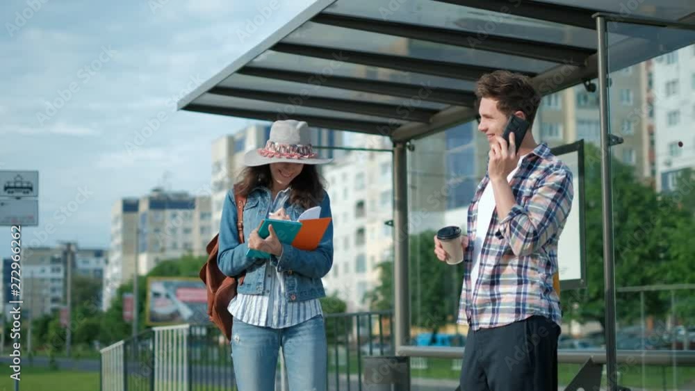 Young man and girl. Standing on the Bus Stop. Sun is Shining. Man Using his Mobile Phone. Waiting for his Tram. City Lifestyle. Young man and girl. Standing on the Bus Stop. Sun is Shining. Man Using his Mobile Phone. Waiting for his Tram. City Lifestyle.