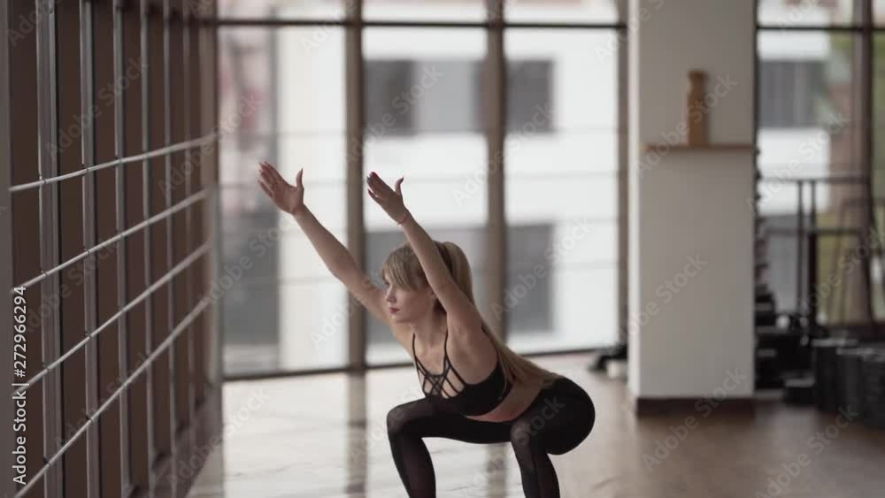Young beautiful slender woman in a fitness room with a glass wall is doing a squatting. Young beautiful slender woman in a fitness room with a glass wall is doing a squatting.