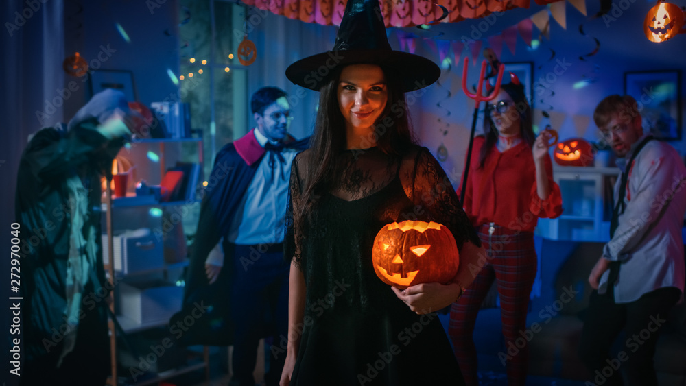 Halloween Costume Party: Gorgeous Seductive Witch Wearing Dress Holds Burning Pumpkin. Background: Beautiful Devil, Scary Death, Count Dracula, Zombie Dancing in the Decorated Room