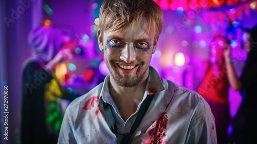 Halloween Costume Party: Portrait of Brain Dead Zombie Wearing Bloody Suit Smiles Creepily. In the Background Monsters Having Fun and Dancing in the Decorated Room © Gorodenkoff