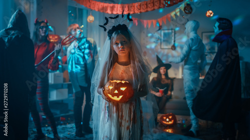 Halloween Costume Party: Little Girl in a Bloody White Bride Dress Holding Scary Doll. Zombie, Blood Thirsty Dracula, Mummy, Bewitching Witch and Dazzling She Devil Having Fun in Decorated Room