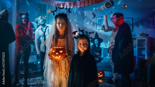 Halloween Costume Party:Portrait Girl in a Bloody White Bride Dress Holding Scary Doll Standing with Cute Little Vampire Bat Sister. In the Background Group of Monsters Dancing and Having Fun 