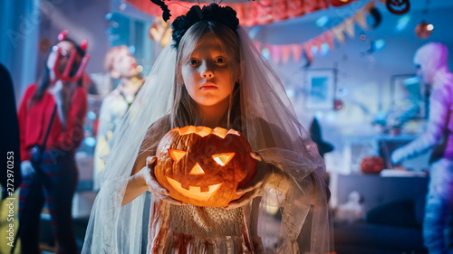 Halloween Costume Party: Smiling Little Girl in a Bloody White Bride Dress Holding Burning Pumpkin Head. Zombie, Blood Thirsty Dracula, Mummy, Bewitching Witch and Dazzling She Devil Dance