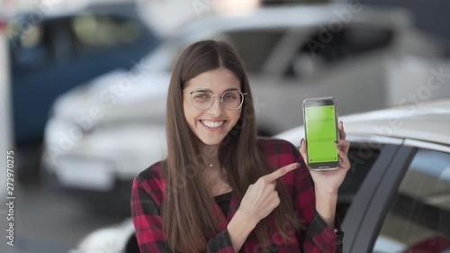 Beutiful young woman is standing on a car show, holding a phone, smiling and demonstrated a green screen of the phone. The screen is green so it could be different advertising there. photo
