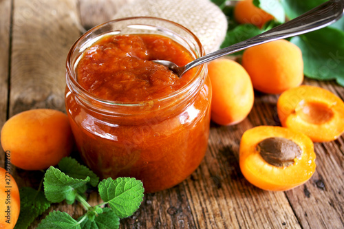 Glass jar of apricot jam on wooden table with ripe apricots at background