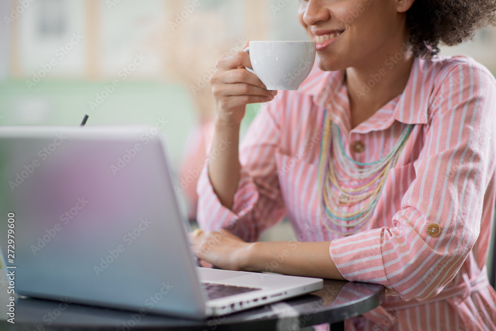 Charming mixed race woman in striped pink dress aitting in coffee shop, drinking coffee and using laptop. Charming mixed race woman in striped pink dress aitting in coffee shop, drinking coffee and using laptop.