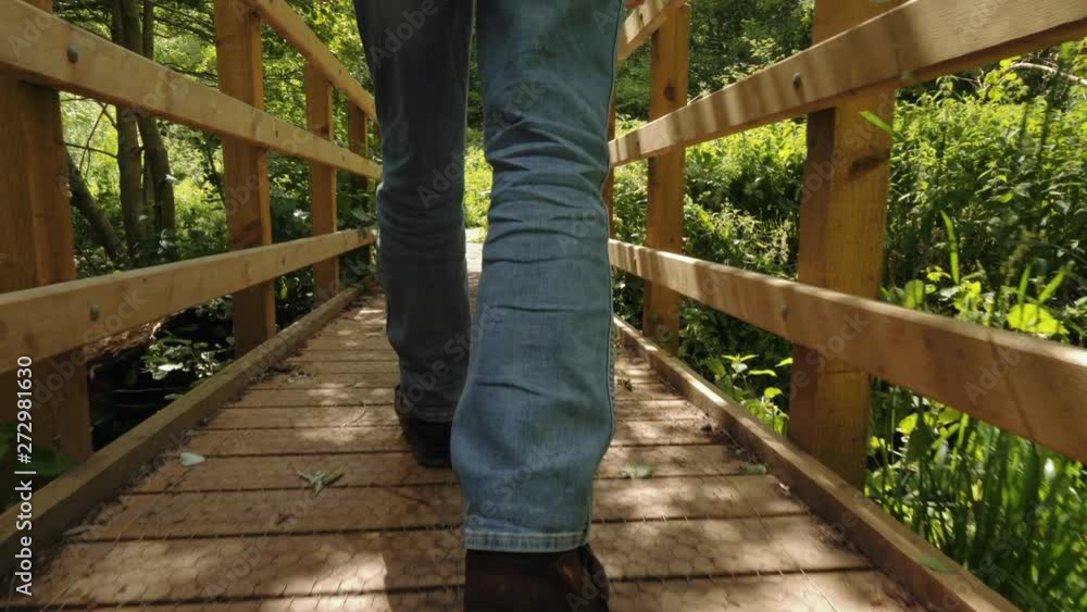 Part 1 - Man (lower half) walking along wooden bridge with sun in front with long shadows. Greenery either side of the wooden path. Part 1 - Man (lower half) walking along wooden bridge with sun in front with long shadows. Greenery either side of the wooden path.