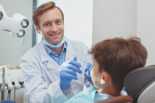 Experienced dentist concept. Handsome mature male dentist smiling joyfully to the camera  working at his clinic  examining teeth of his little patient