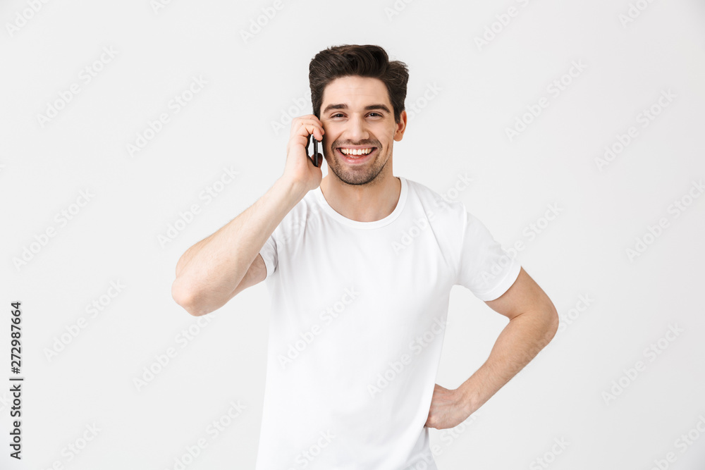 Happy young man posing isolated over white wall background talking by mobile phone. Happy young man posing isolated over white wall background talking by mobile phone.