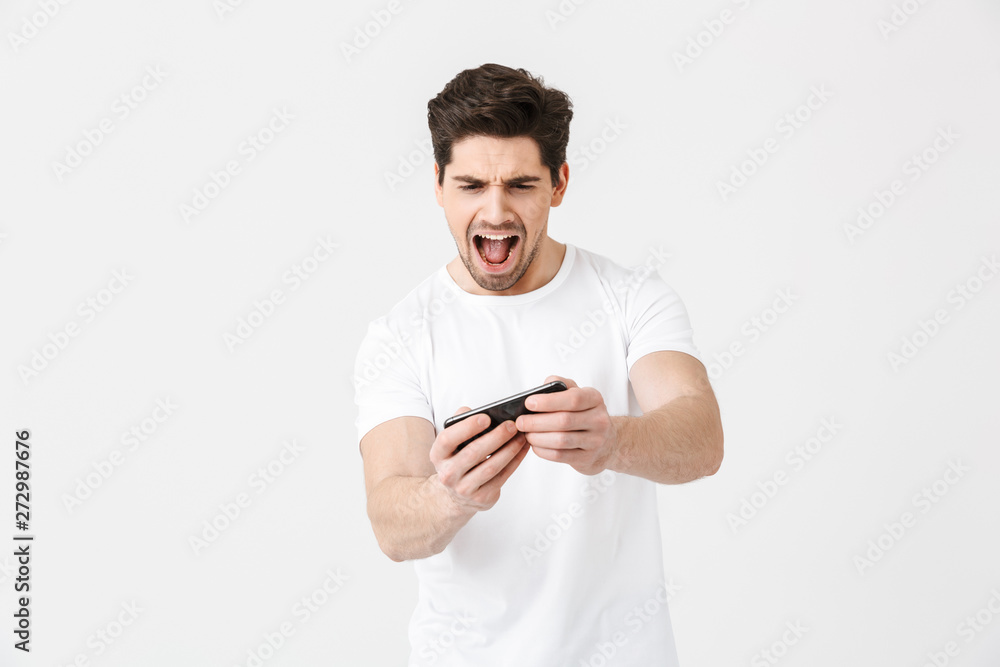 Excited nervous emotional young man posing isolated over white wall background play games by mobile phone. Excited nervous emotional young man posing isolated over white wall background play games by mobile phone.