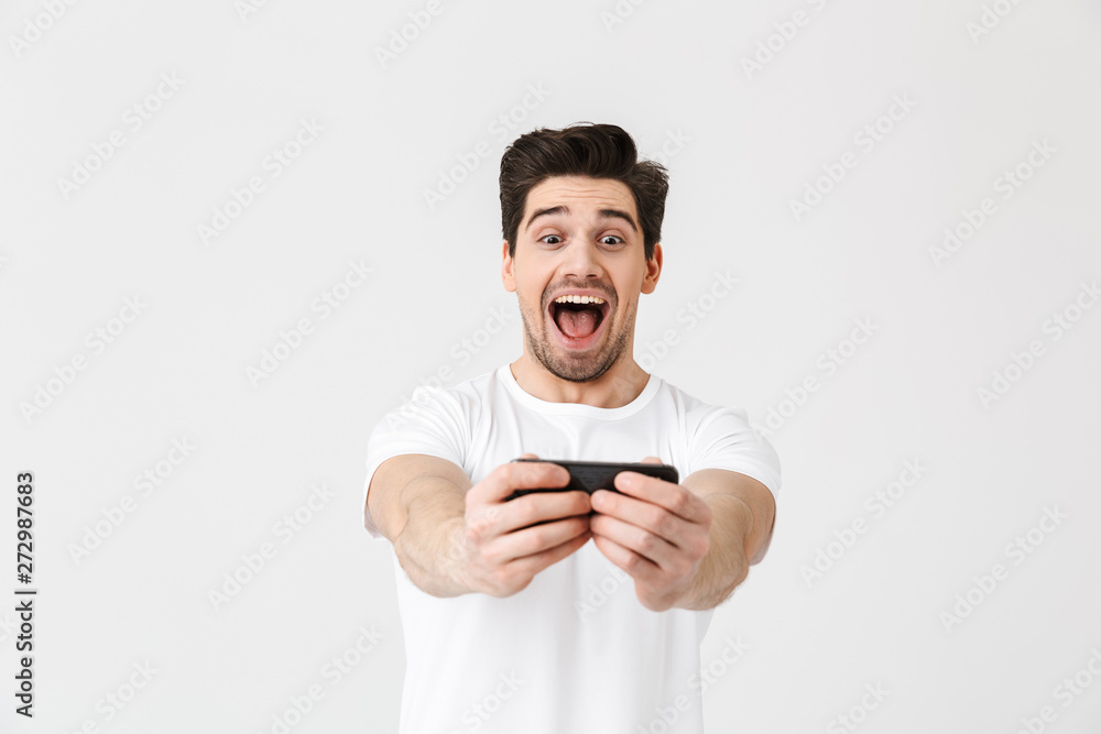 Excited happy emotional young man posing isolated over white wall background play games by mobile phone. Excited happy emotional young man posing isolated over white wall background play games by mobile phone.
