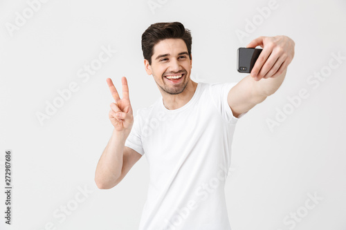 Emotional young man posing isolated over white wall background showing peace gesture take selfie by mobile phone.