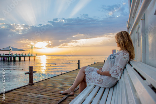 A girl in a beach tunic meets dawn on a bench near the sea. © Sergey