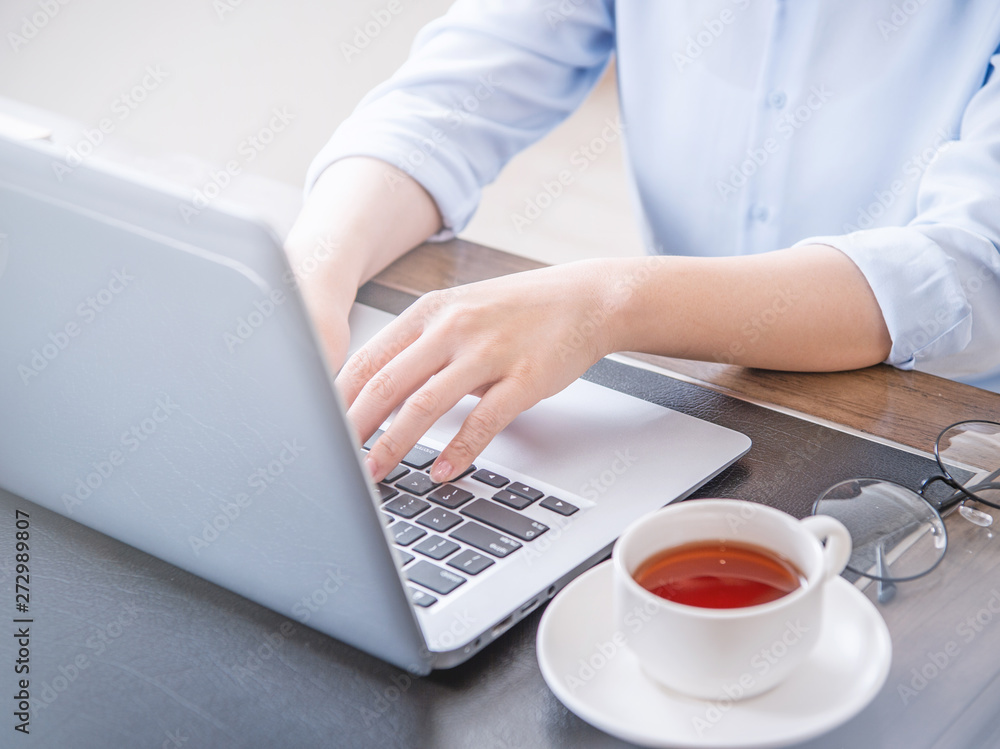 Business concept. Woman in blue shirt typing on computer with coffee on office table, backlighting, sun glare effect, close up, side view, copy space Business concept. Woman in blue shirt typing on computer with coffee on office table, backlighting, sun glare effect, close up, side view, copy space