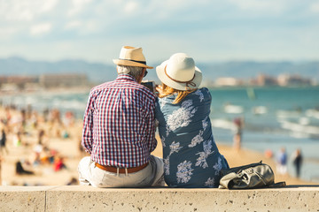 Loving the elderly couple sitting on the wall facing the beach, watching and taking pictures of the landscape on a romantic trip