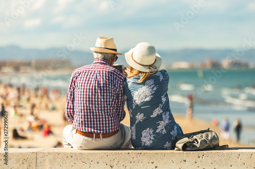 Loving the elderly couple sitting on the wall facing the beach, watching and taking pictures of the landscape on a romantic trip