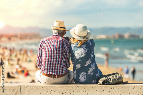 Mature couple of retired lovers enjoying retirement on the beach facing the sea with mobile cell phone taking pictures at sunset. Couple happy true love in the nature