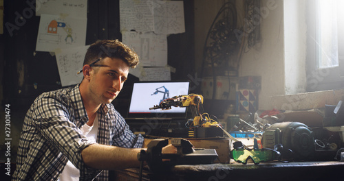 Portrait of an young professional engineer is smiling in camera during his work on projection of an innovative technology mechanical arm in his workshop.