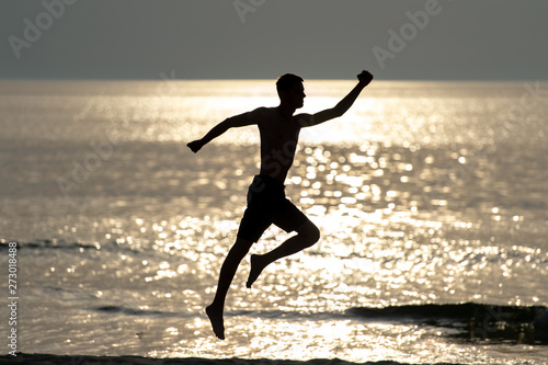 Dark silhoutte of man jumping with hands apart like superhero over sea with sun light on background. Unrecognizable man dancing beyond sea at sunset in summer evening. Freedom and happiness concept.