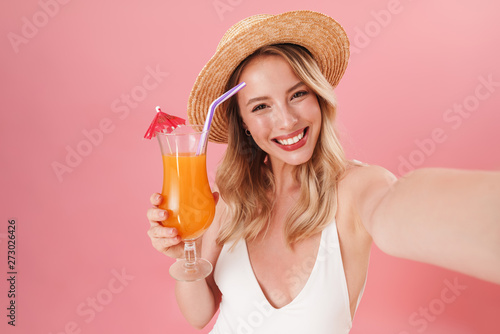 Excited young woman posing isolated over pink wall background dressed in swimwear beach concept drinking cocktail take selfie by camera.