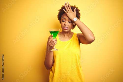 Young african american woman with afro hair drinking a cocktail over yellow isolated background stressed with hand on head, shocked with shame and surprise face, angry and frustrated. Fear and upset