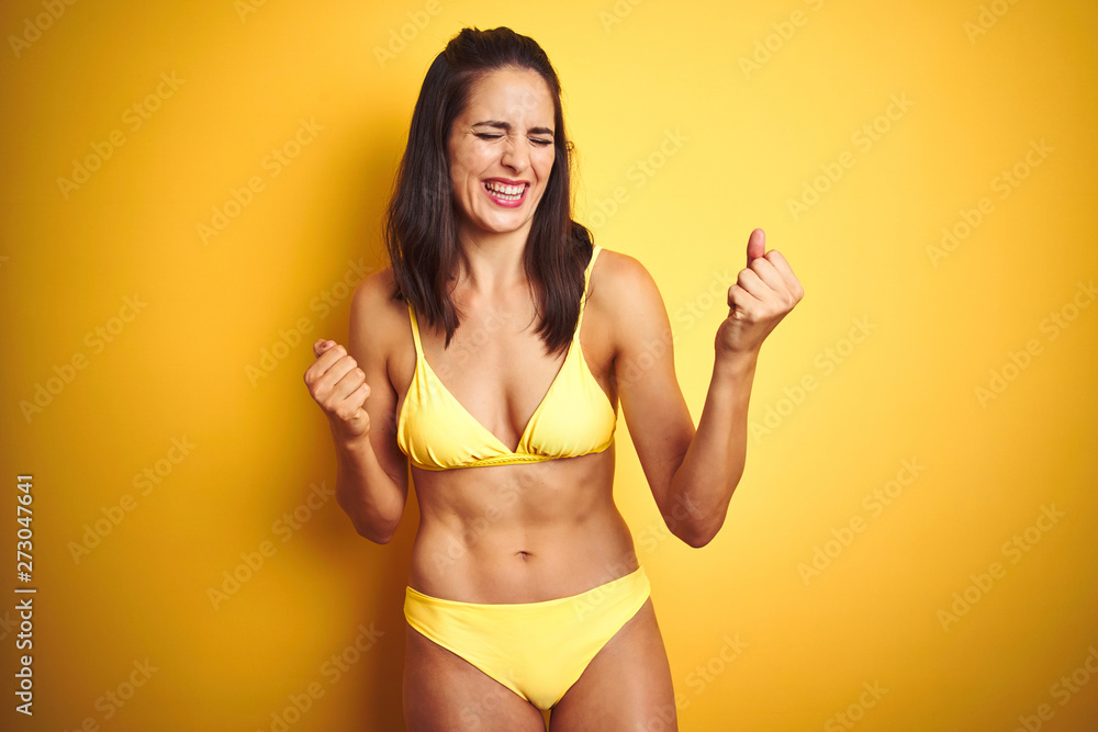 Beautiful woman wearing yellow bikini on summer over isolated yellow background very happy and excited doing winner gesture with arms raised, smiling and screaming for success. Celebration concept. Beautiful woman wearing yellow bikini on summer over isolated yellow background very happy and excited doing winner gesture with arms raised, smiling and screaming for success. Celebration concept.
