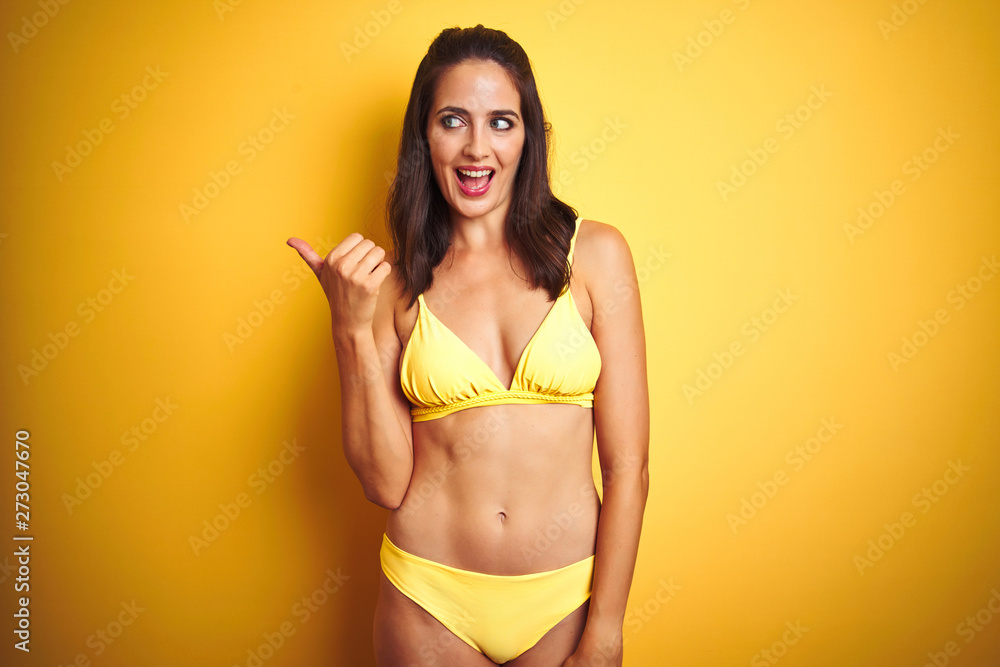 Beautiful woman wearing yellow bikini on summer over isolated yellow background smiling with happy face looking and pointing to the side with thumb up. Beautiful woman wearing yellow bikini on summer over isolated yellow background smiling with happy face looking and pointing to the side with thumb up.