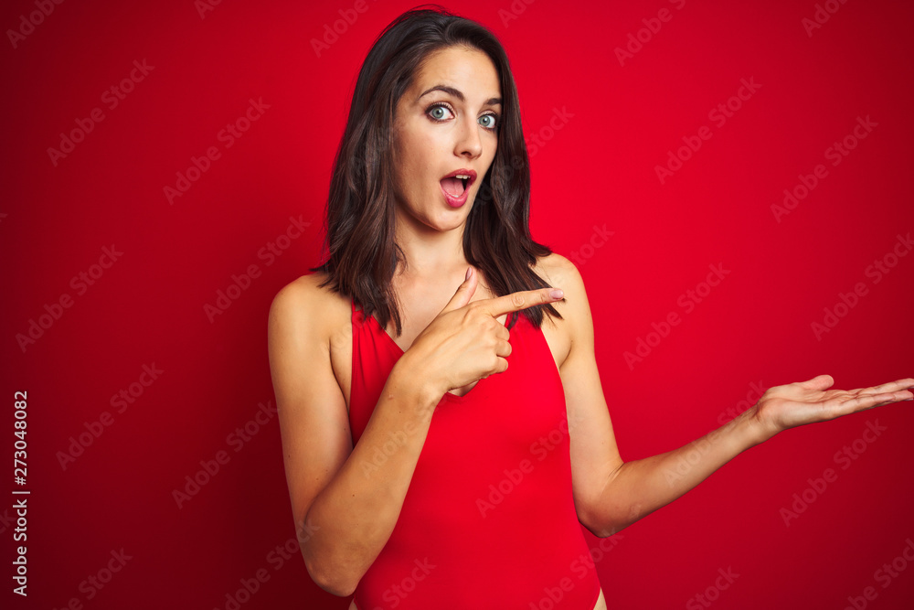Beautiful woman wearing bikini swimwear over red isolated background amazed and smiling to the camera while presenting with hand and pointing with finger. Beautiful woman wearing bikini swimwear over red isolated background amazed and smiling to the camera while presenting with hand and pointing with finger.