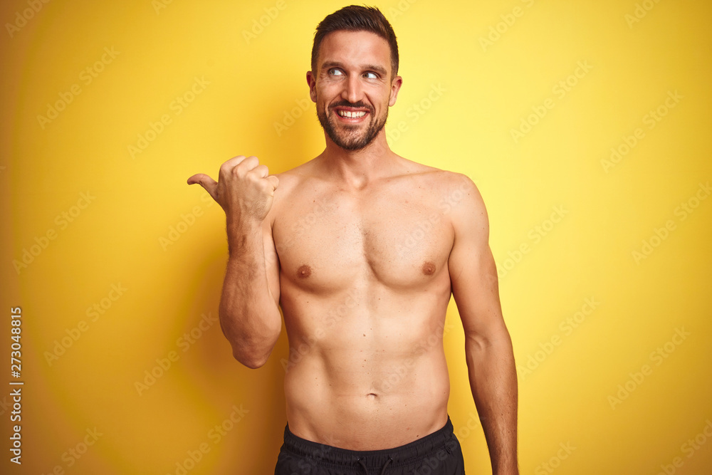 Young handsome shirtless man over isolated yellow background smiling with happy face looking and pointing to the side with thumb up.